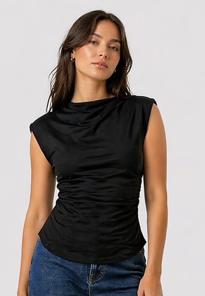 Mujer con cabello largo y castaño que lleva una blusa negra sin mangas con fruncidos y vaqueros azules sobre un fondo claro y liso.