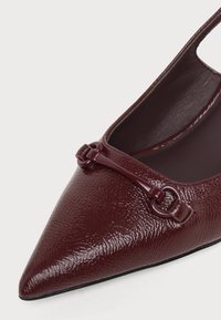 Chaussure plate pointue rouge foncé brillante avec surface texturée et détail décoratif en barre sur le devant.