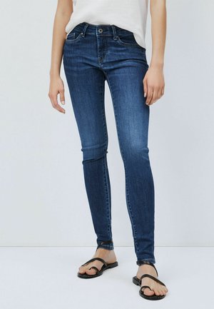 Vrouw draagt donkerblauwe skinny jeans en zwarte sandalen met bandjes en platte zolen, staand tegen een effen witte achtergrond.