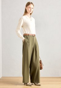 Beżowy sweter z dzianiny z kołnierzykiem, w zestawieniu z oliwkowo-zielonymi spodniami o wysokim stanie i szerokich nogawkach, oraz jasnobrązowym paskiem. Brązowa torebka jako akcent.