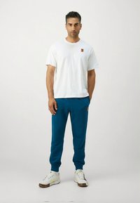 Bela majica s kratkimi rokavi in zeleno-modre športne hlače. Majica ima majhen logotip; hlače so z bočnimi žepi in zoženim krojem. Bela športna obutev.