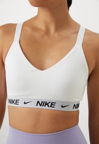 Hvit sports-BH med V-hals design og justerbare stropper. Har et grått elastisk bånd med svart "Nike"-logo. Glatt tekstur.