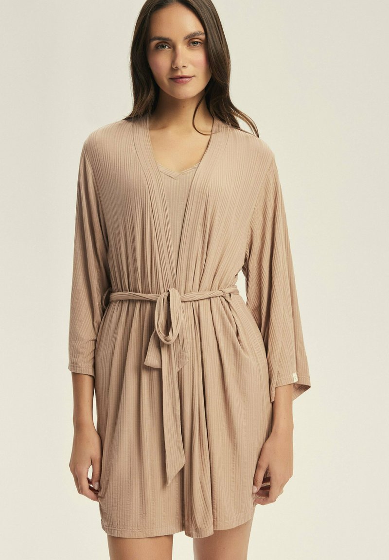 Beżowa sukienka w prążki z długimi rękawami, dekoltem w serek i przewiązanym paskiem w talii. Ma luźny fason i miękką teksturę.