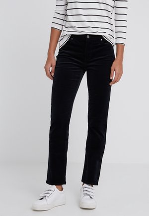 Personne portant un pantalon noir à jambe droite, des baskets blanches et une chemise blanche à manches longues avec des rayures horizontales noires.