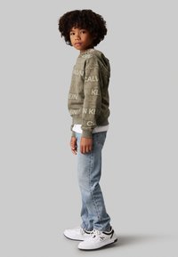 Felpa con cappuccio verde oliva e stampa bianca del logo, abbinata a jeans azzurri chiari e sneakers bianche. Design semplice, vestibilità comoda.