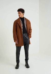 Casaco castanho texturizado com gola lapela, fecho de botão, sobre uma camisa cinza e um suéter de gola alta negro, combinado com calças pretas e botas.
