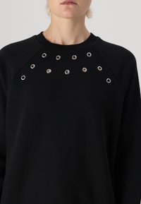 Sudadera negra con ojales plateados dispuestos en una fila a lo largo del pecho. Hecha de una tela suave con un cuello redondo clásico y mangas raglán.