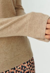 Maglione beige lavorato a maglia con polsini a costine, texture morbida e vestibilità leggermente ampia. Gonna patterned con motivi floreali visibili nella parte inferiore.