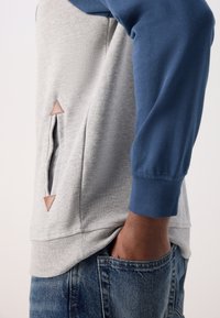 Personne portant une chemise à manches longues grise et bleue, la main dans la poche, montrant un détail de poche latérale avec deux petits accents triangulaires marron.