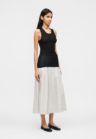 Λεπτή γυναίκα που στέκεται φορώντας μαύρο τοπ χωρίς μανίκια, μίντι φούστα σε ανοιχτό γκρι χρώμα με πιέτες και μαύρα φλατ παπούτσια σ ε λευκό φόντο.