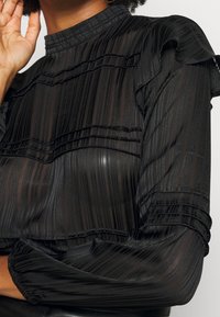 Haut noir transparent avec des rayures verticales texturées. Présente des manches à volants et un col montant, mettant en valeur des détails de tissu superposés.