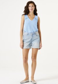 Hellblauses ärmelloses Strickoberteil mit V-Ausschnitt und Spitzenakzenten, kombiniert mit hellen Jeansshorts mit umgeschlagenem Saum und flachen Schuhen.