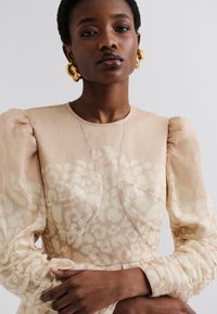 Blusa beige con maniche a sbuffo, tessuto testurizzato e leggeri motivi astratti. Presenta polsini arricciati e dettagli di cucitura sottili.