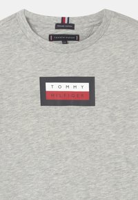 Šedé tričko z organické bavlny s obdélníkovým logem, na kterém je "TOMMY" napsáno bílou barvou a "HILFIGER" černou barvou na červeném pozadí. Krátké rukávy.