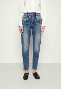 Jeans de ganga azul com um corte slim, cinco bolsos e desbotamento subtil. Usados com uma camisa branca de botão e sapatos pretos tipo loafers.