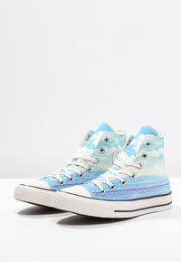 Vysoce střižené plátěné tenisky s bílými tkaničkami a potiskem modré oblohy a mraků na textilu, umístěné vedle sebe na bílé ploše.