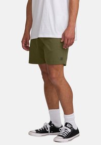 Pantalones cortos de color verde oliva con una textura suave, bolsillos laterales y un pequeño parche con logo. Combinados con una camiseta blanca y zapatillas negras.
