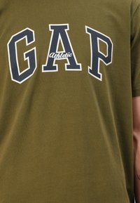 Olivenfarbiges Baumwoll-T-Shirt mit auffälligem weißen und marineblauen "GAP Athletic"-Schriftzug. Kurze Ärmel, runder Hals, glatte Textur.