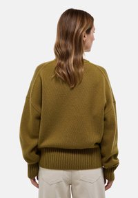 Pull oversized vert olive avec un ourlet et des poignets côtelés, tricot texturé et épaules tombantes, porté avec un pantalon de couleur claire.