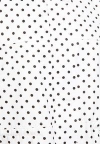 Hvidt stof med jævnt fordelte små sorte polkaprikker og synlige sømme, der viser et nærbillede af tekstur og mønster på et tøjstykke.