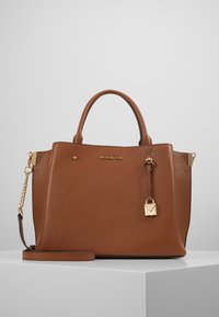 Brązowa skórzana torebka na ramię z górnymi uchwytami, odczepianym paskiem na ramię, złotym łańcuszkiem oraz zawieszką w kształcie kłódki na białej powierzchni.