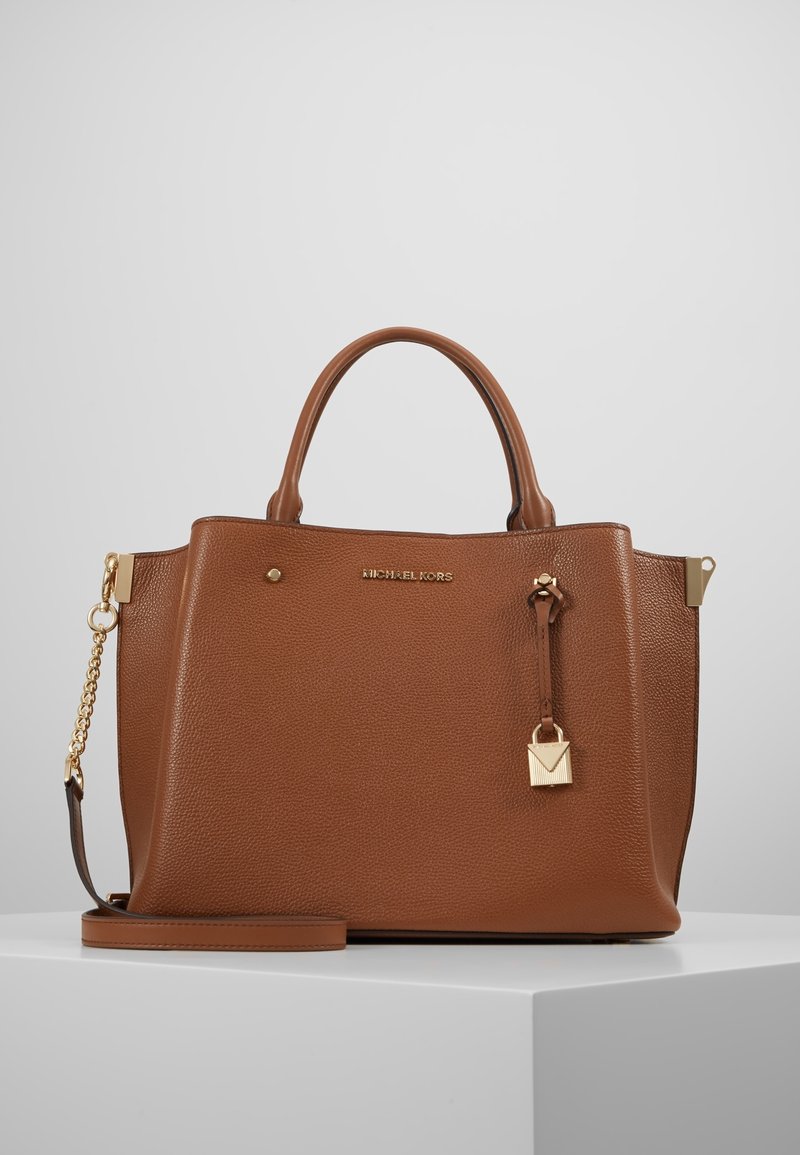 Brązowa skórzana torebka na ramię z górnymi uchwytami, odczepianym paskiem na ramię, z�łotym łańcuszkiem oraz zawieszką w kształcie kłódki na białej powierzchni.