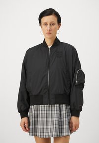 Black bomber jacket with a zip front, ribbed hem, and cuffs; features an embroidered logo and a zip pocket on the left sleeve.