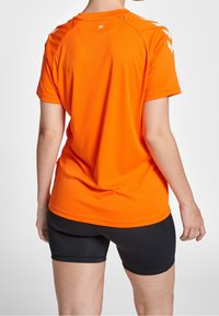 Koszulka sportowa z krótkim rękawem w jasnopomarańczowym kolorze o gładkiej fakturze, z białymi akcentami na ramionach i logo na karku z tyłu.