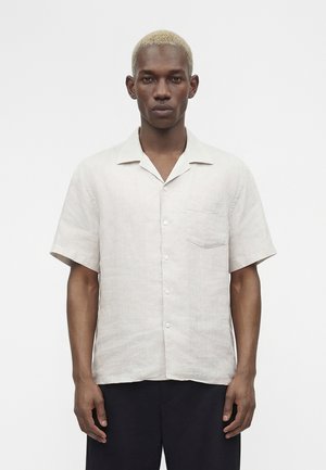 Mann med kort blond hår iført en lys beige kortermet skjorte med knapper og mørke bukser, stående mot en ensfarget hvit bakgrunn.