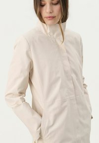 Ljust beige, skräddarsydd kavaj med hög krage, lång ärm och sidofickor. Tyget ser slätt ut med minimal sömdetalj.