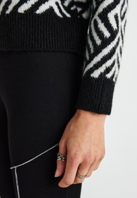 Pull en maille noire avec des motifs géométriques blancs, manches à finitions côtelées ; associé à un pantalon noir à côtes ; main ornée de bagues en argent.