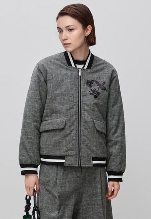 Young person wearing gray plaid jacket and matching pants, holding a black twisted handle bag, looking down and to the side.