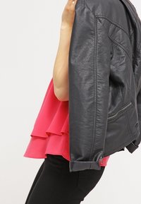 Chaqueta de cuero negra con un acabado texturizado, sostenida junto a una blusa peplum de capas en un brillante tono rosa. Pantalones ajustados negros completan el look.