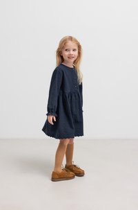 Lächelndes Mädchen mit langen blonden Haaren trägt ein dunkles Kleid und kniehohe Socken, steht auf einem hellen Boden vor einem schlichten Hintergrund.