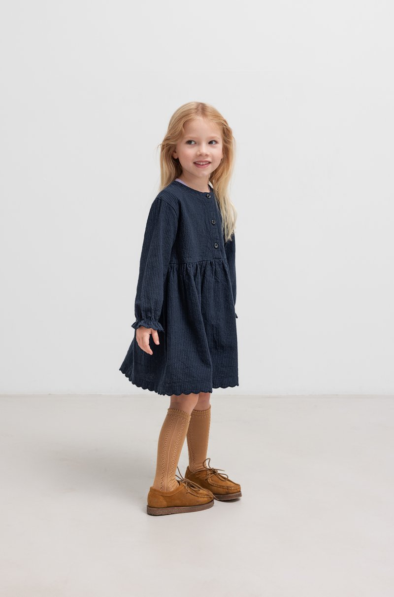 Lächelndes Mädchen mit langen blonden Haaren trägt ein dunkles Kleid und kniehohe Socken, steht auf einem hellen Boden vor einem schlichten Hintergrund.