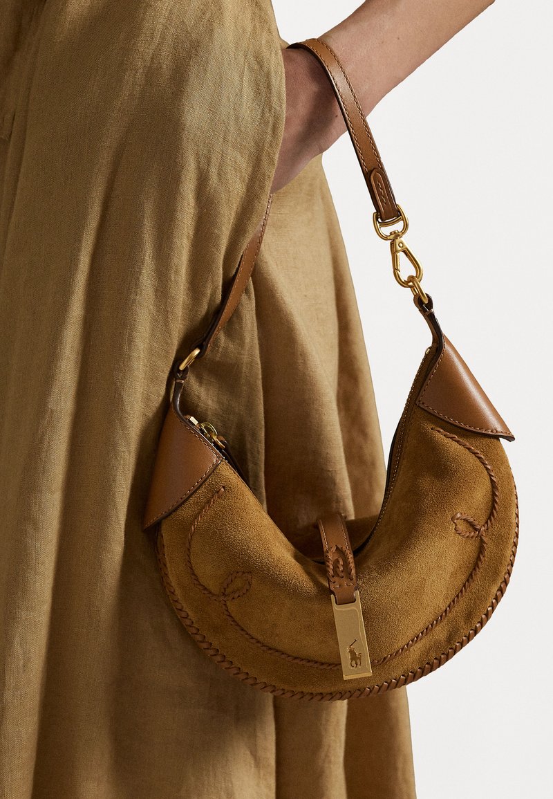 Mala de camurça castanha com toques de couro, em forma de crescente, com costura detalhada e acessórios em dourado. Alça ajustável, apoiada contra um tecido texturizado.