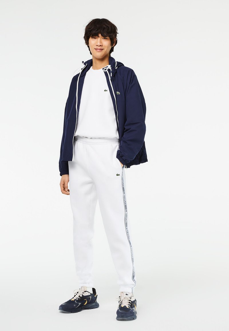 Veste zippée navy avec capuche, tee-shirt blanc à col rond, sweatpants blancs avec des bandes latérales, et baskets navy avec des accents beiges.