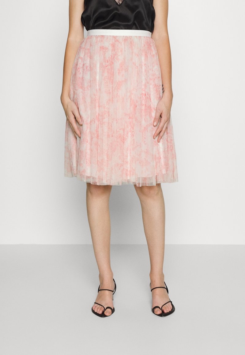 Femme portant une jupe en tulle rose plissée longueur genou avec ceinture blanche, haut noir sans manches et sandales noires à brides, debout contre un mur blanc.