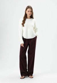 Pull blanc en maille à encolure ronde associé à un pantalon ample bordeaux. Le modèle porte des sandales noires, affichant un look élégant et moderne.