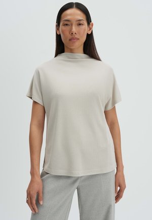 Donna che indossa una maglietta a maniche corte di colore beige chiaro con collo alto abbinata a pantaloni grigi larghi su sfondo neutro.