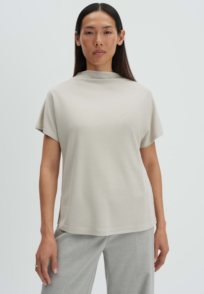 Donna che indossa una maglietta a maniche corte di colore beige chiaro con collo alto abbinata a pantaloni grigi larghi su sfondo neutro.