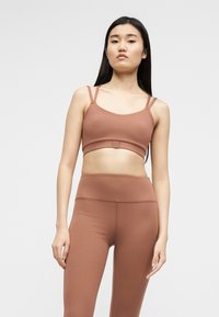 Νεαρή γυναίκα με μακριά μαύρα μαλλιά φοράει καφέ αθλητικό τοπ και ψηλόμεσα κολάν, στέκεται μπροστά σε ένα απλό φωτεινό φόντο.