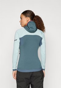 Kapuutsiga jakk, millel on teal värvi keha, heledamad varrukad, kontrastne tume õlavarre paneel ja roosa aktsent kapuutsis.