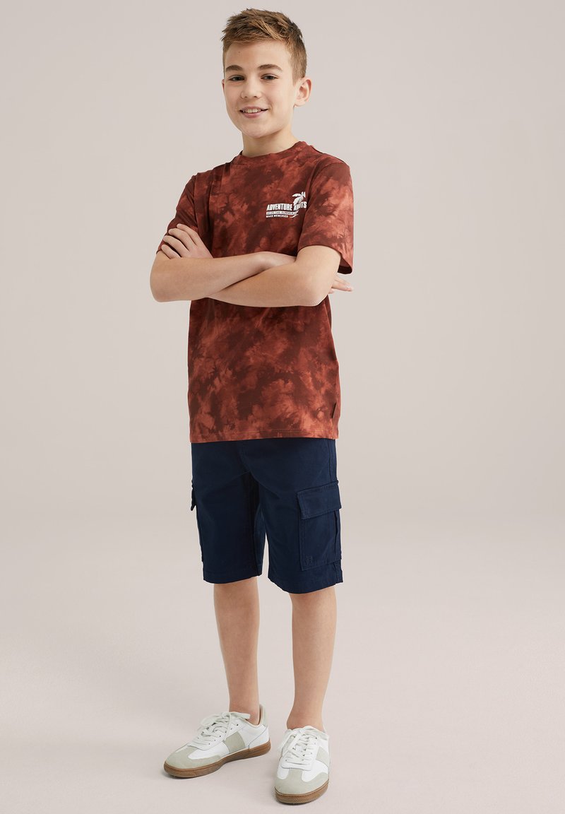 Czerwony t-shirt z krótkim rękawem w stylu tie-dye, granatowe szorty cargo i białe trampki z szarymi akcentami i brązowymi podeszwami. Swobodny strój na ciepłe dni.