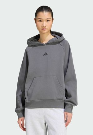 Vrouw draagt een losse grijze Adidas hoodie met voorzak en witte joggingbroek, staand tegen een eenvoudige lichte achtergrond.