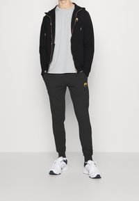 Černá mikina na zip, světle šedé tričko, tmavě šedé tepláky s oranžovým logem a bílé tenisky s námořnickými akcenty. Jednoduchý sportovní design.