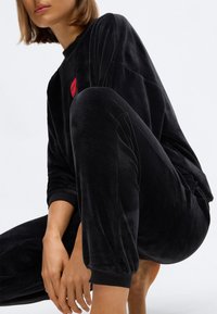 Zwarte fluwelen loungewear set met een sweatshirt met ronde hals en een rood logo, en bijpassende broek met elastische manchetten en een gladde textuur.