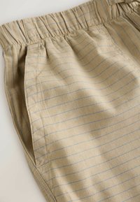 Pantalon en tissu beige avec de fines rayures bleues horizontales, une taille élastique et des poches latérales visibles sur une surface plane.