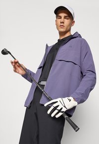 Lilla golfjakke med glidelås foran, justerbar hette, båret over en svart polo. Modellen holder en golfkølle med en hvit hanske med svarte detaljer.