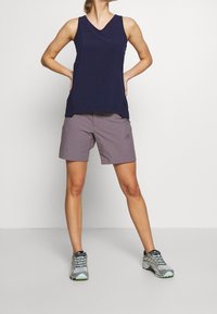 Marinblus utan ärmar med V-hals; matchad med ljuslila shorts. Modellen bär grå sportskor med turkosa accenter.